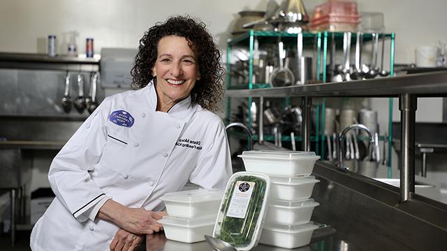 Diane Mora, wearing a white chef’s jacket that reads “Diane Mora, Kids Feeding Kids,” stands next to a metal culinary counter with her arm propped on food container boxed used in the Kids Feeding Kids program.