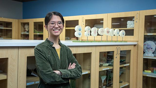 Biology tutor Kaitlyn Sy stands in a science lab room with cabinets containing scientific teaching tools around her. To her right is a display showing how a cell divides.  